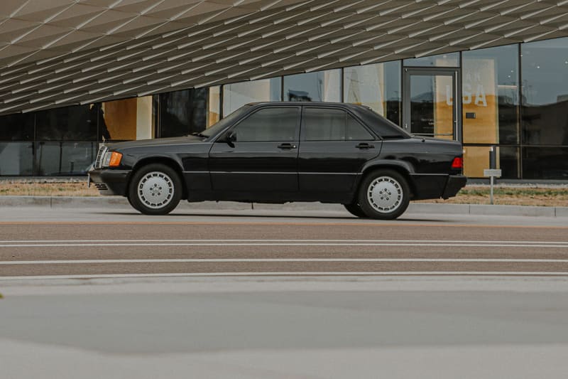 A black Mercedes-Benz 190E parked small in the frame beneath a vast pale coffered concrete canopy.