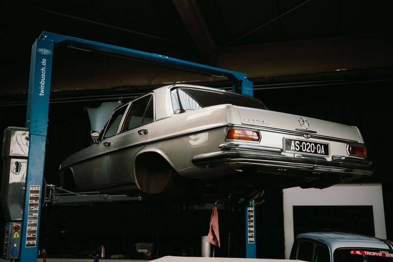 A silver 1970s Mercedes-Benz sedan raised on a two-post lift in a dark shop.