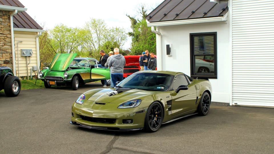 A small morning meet on a pale asphalt lot: a green sports car in front, an older one with its hood up, and 1960s coupes behind.