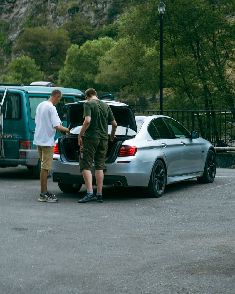 Two people seen from behind at the open trunk of a silver sedan in a mountain parking area.