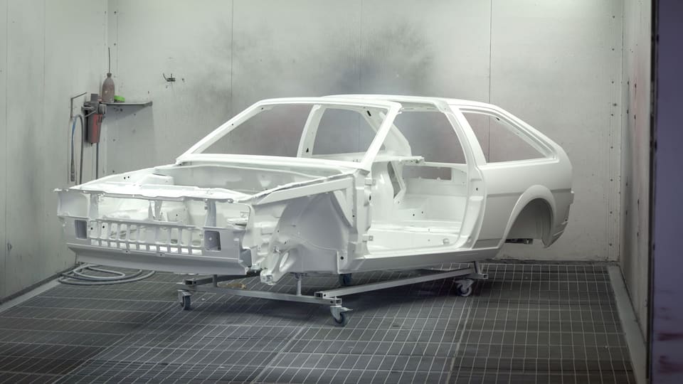 A white primered hatchback shell on a dolly in a spray booth.