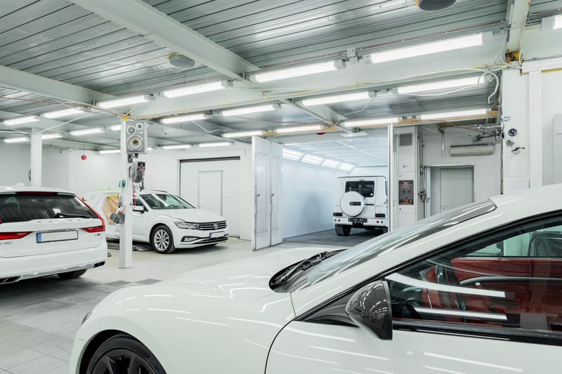 A bright white service hall with several late-model cars in adjacent bays and a paint booth at the far end.