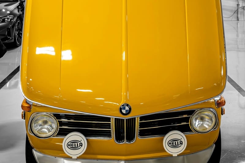 A yellow 1970s sedan photographed head-on with the hood raised in a white showroom.